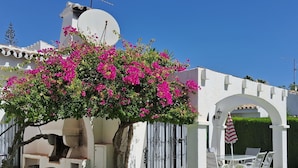 Edifici blanc amb una antena parabòlica, una pergola amb flors roses i un pati amb taula i cadires.
