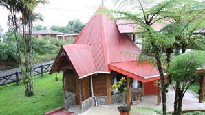 Una pequeña estructura de madera con techo rojo y base de piedra, rodeada de abundante follaje y un sendero pavimentado.