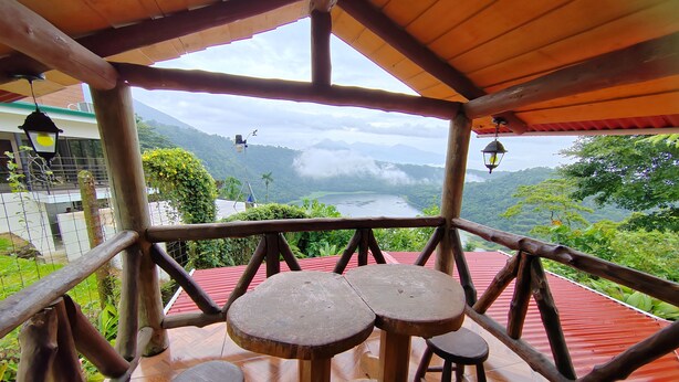 Una terraza de madera con una mesa y taburetes, con vistas a un paisaje pintoresco con montañas y un cuerpo de agua.