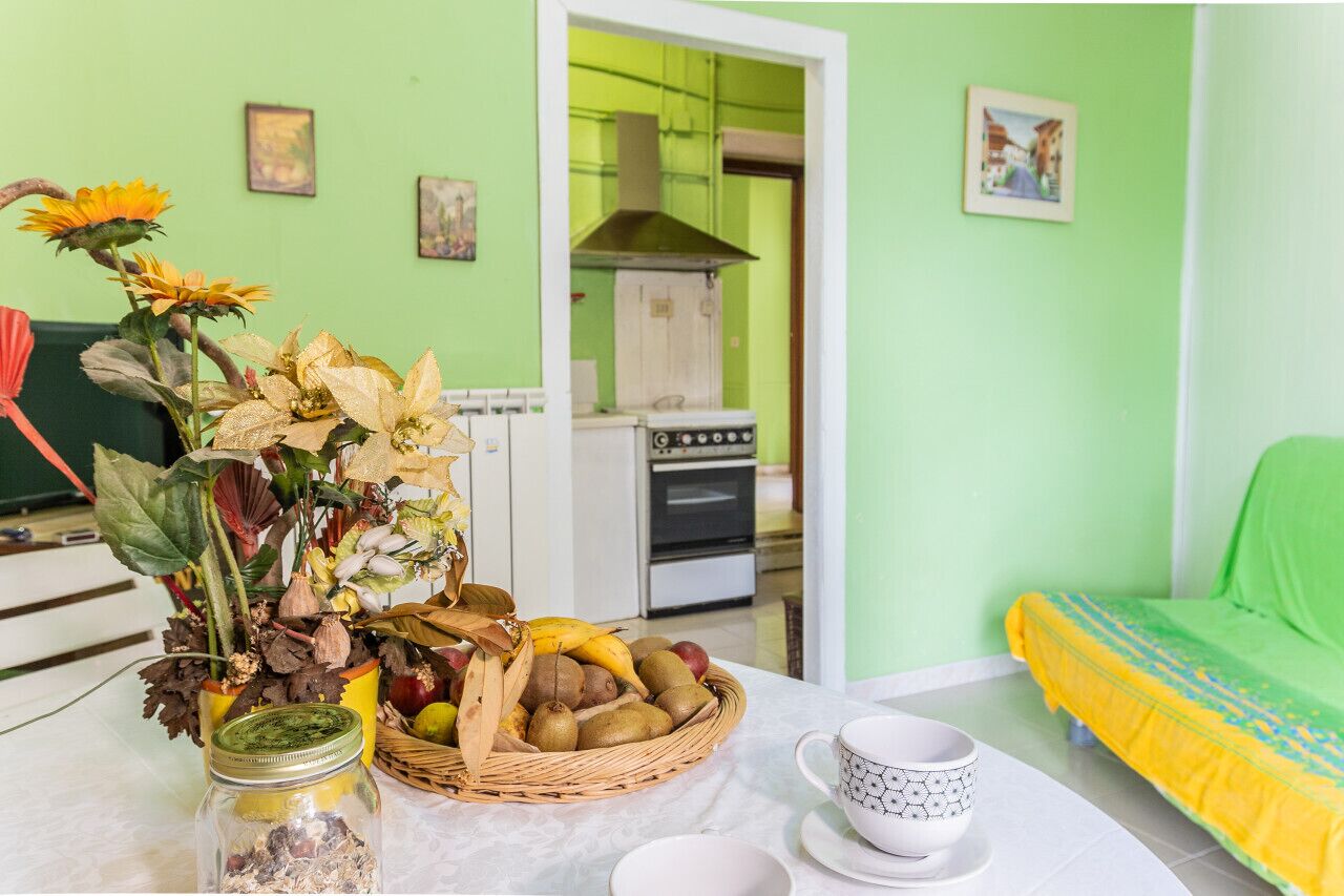 Een kamer met een groene wand, een keuken met een kookplaat en oven, een eettafel met een mand vol fruit en een vaas met gedroogde bloemen, en een bed met een gele en blauwe sprei.