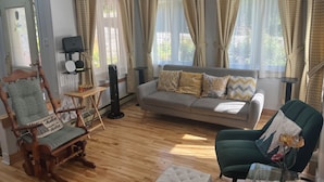 Un salon avec un canapé, un fauteuil à bascule et un fauteuil vert. On y trouve une petite table avec un miroir et un haut-parleur.