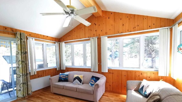 Une pièce chaleureuse avec des murs en bois, un ventilateur de plafond, un canapé beige et une fenêtre avec des rideaux.