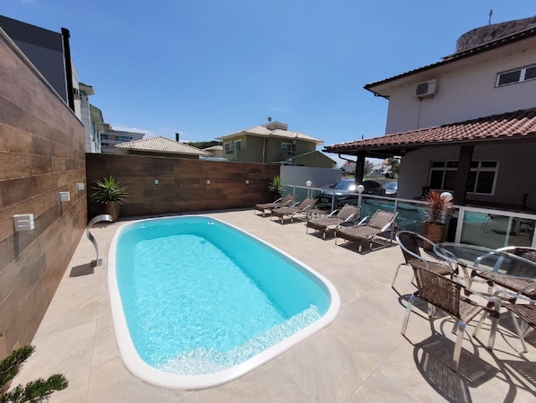 A rectangular swimming pool with a tiled deck, surrounded by lounge chairs and a glass table.