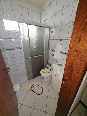 A bathroom with white tiled walls, a shower area, a toilet with a decorative cover, and a sink with a green soap dish.