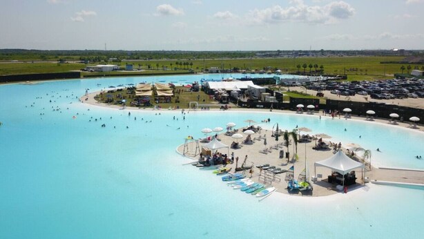 En stor udendørs swimmingpool med flere baner, en central ø med en legekonstruktion, og et område omkring med parasoller og liggestole.