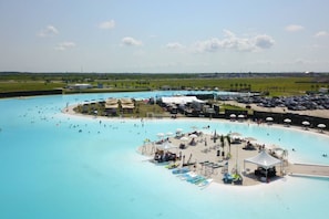A large outdoor swimming pool with multiple lanes, a central island with a play structure, and a surrounding area with umbrellas and lounge chairs.