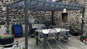 A stone building with a thatched roof, a pergola with a table and chairs underneath, and potted plants around.