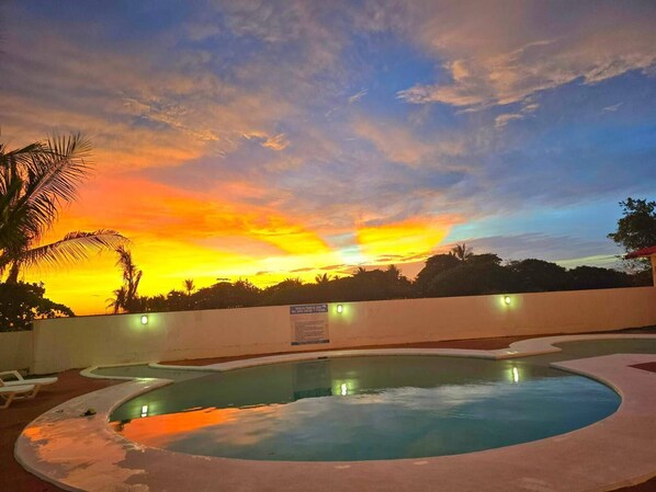 Una piscina con vista al atardecer, una pared blanca y una palmera.