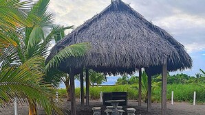 A thatched-roof structure with a bench and a table, surrounded by a palm tree and a pathway leading to it. - Spectacular beachfront apartment, Spacious terrace with an unparalleled view. (Palo Grande)
