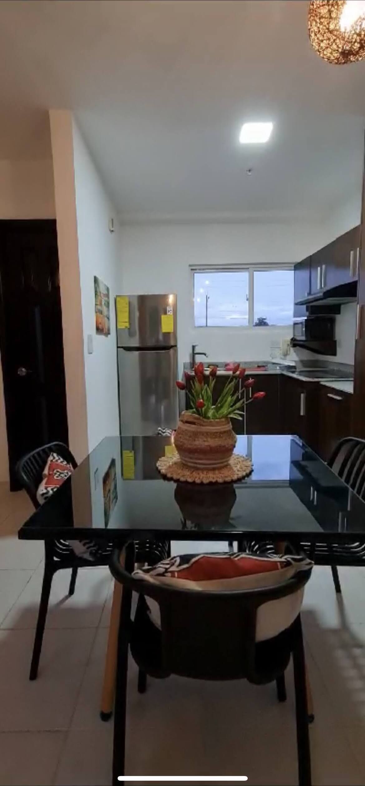 A modern kitchen with a glass dining table, black chairs, a refrigerator, and a window.