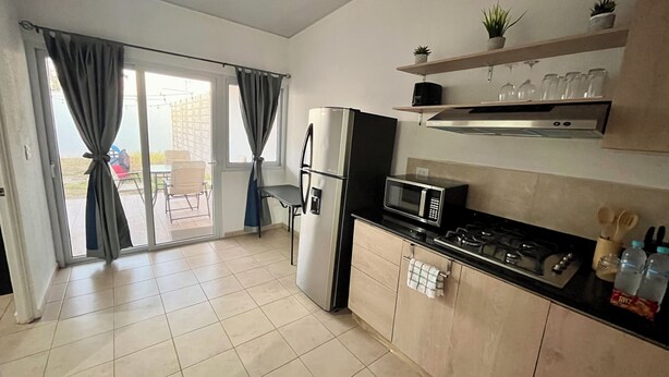 Une cuisine moderne équipée d’un réfrigérateur, d’un four à micro-ondes, d’une cuisinière et de placards. Une porte-fenêtre coulissante donne accès à un espace extérieur aménagé avec une table et des chaises.