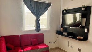 Un salon avec un canapé rouge, un téléviseur fixé au mur, une fenêtre avec un rideau et une petite plante posée sur l’étagère.