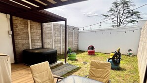 Une terrasse extérieure avec une table et des chaises en verre, un barbecue et un espace de jeux comprenant une balançoire faite d’un pneu et un petit arbre.