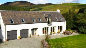 Un bâtiment blanc doté d’un toit en pente, de nombreuses fenêtres et d’une terrasse pavée aménagée avec du mobilier d’extérieur.