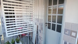 A small, enclosed outdoor area with a white door, a window with blinds, and a white metal fence. There are potted plants on a white bench. - Jardim das Orquídeas 1 (Rio das Ostras)