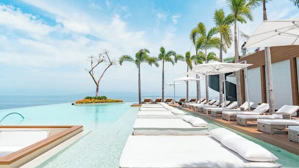 Un espace aménagé au bord de la piscine, avec des chaises longues et des parasols, des palmiers et une vue sur l’océan.
