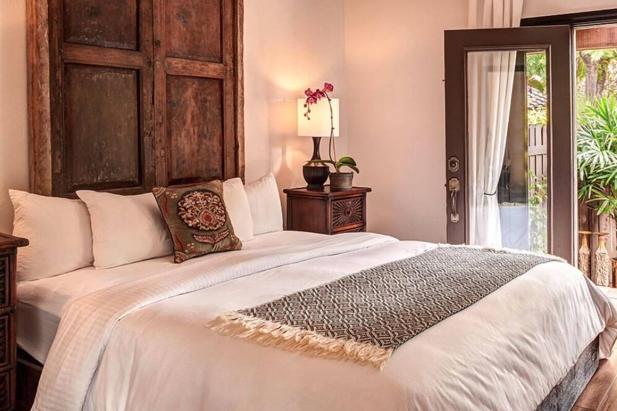 A bedroom with a large bed, a wooden wardrobe, a nightstand with a lamp, and a view of greenery through a door.