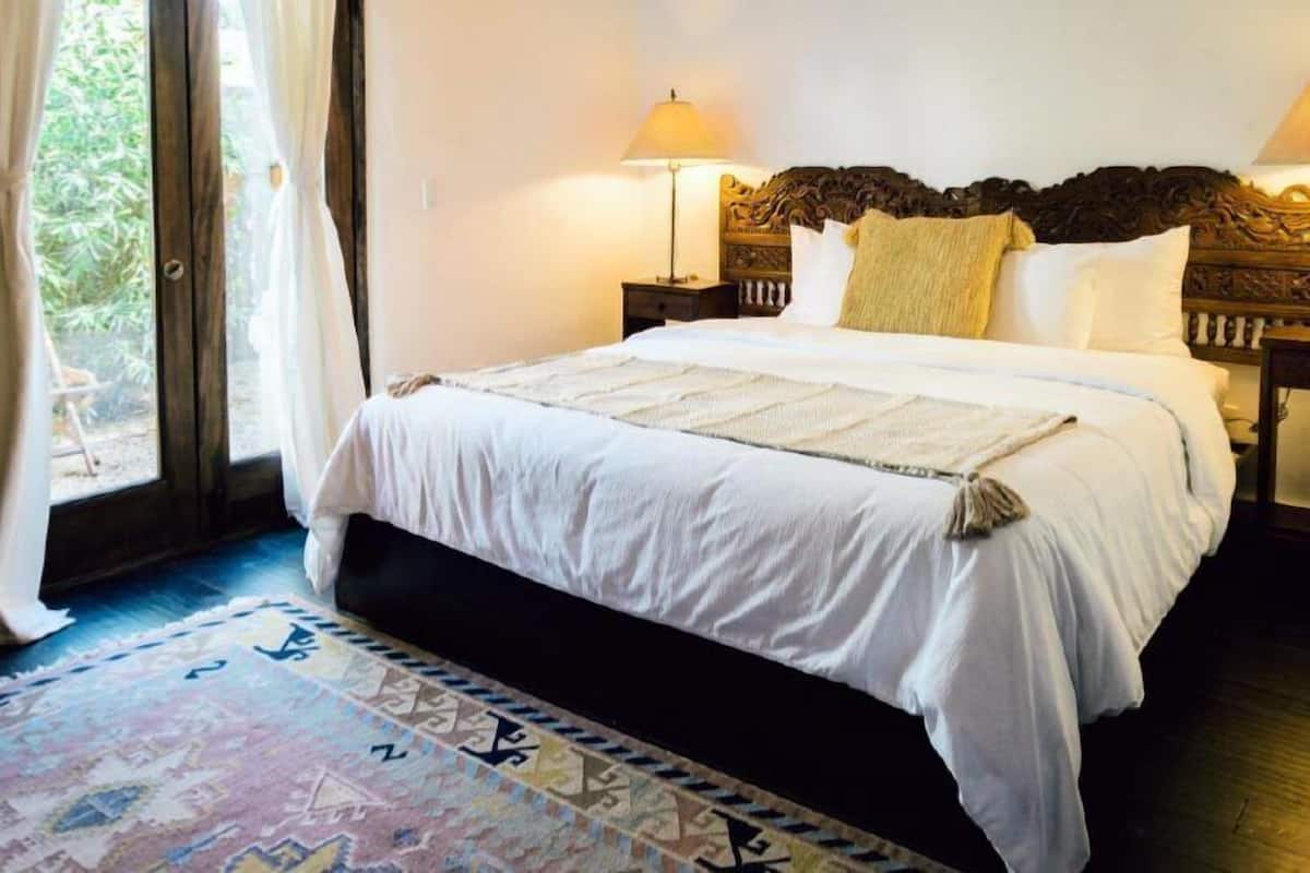 A neatly made bed with a white bedspread and a decorative throw, a wooden headboard, a bedside lamp, and a patterned rug on a wooden floor.