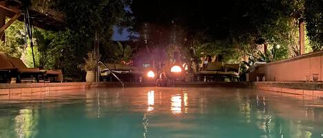 A nighttime pool area with a clear view of the pool's edge, surrounding trees, and a distant illuminated structure.
