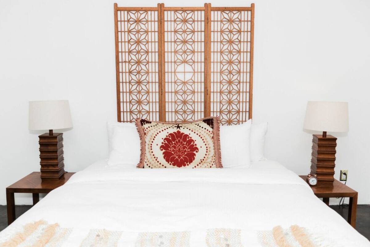 A neatly made bed with a white duvet and a decorative pillow, flanked by two bedside tables with lamps and clocks, against a plain white wall.