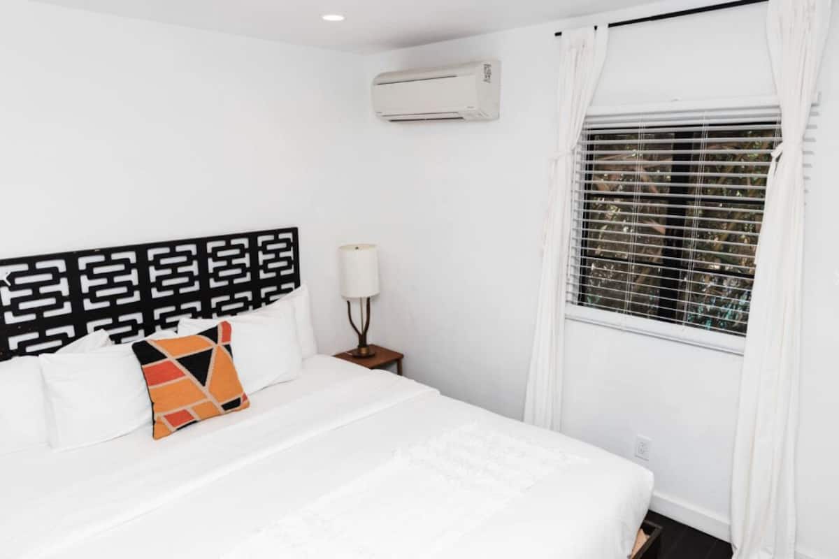 A hotel room with a bed, a nightstand with a lamp, an air conditioning unit, and a window with blinds.