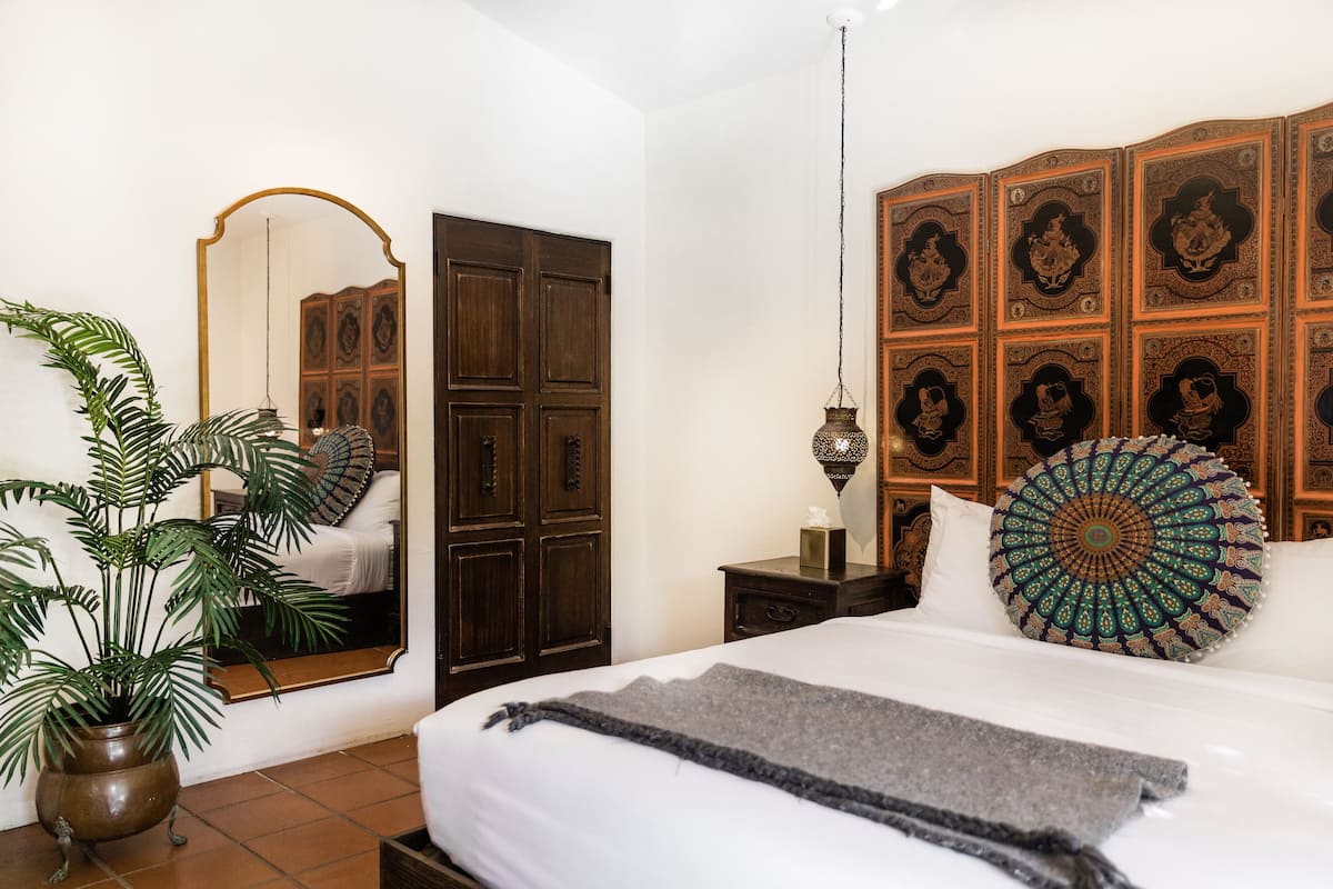 A bedroom with a large bed, a mirror, a potted plant, a wooden wardrobe, and a decorative wall panel.