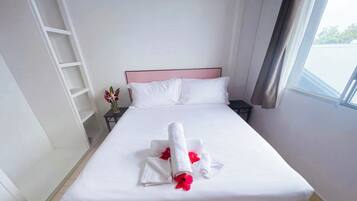 Una cama bien hecha con una toalla blanca enrollada y flores rojas, una mesita de noche con un florero, y una ventana con cortinas.