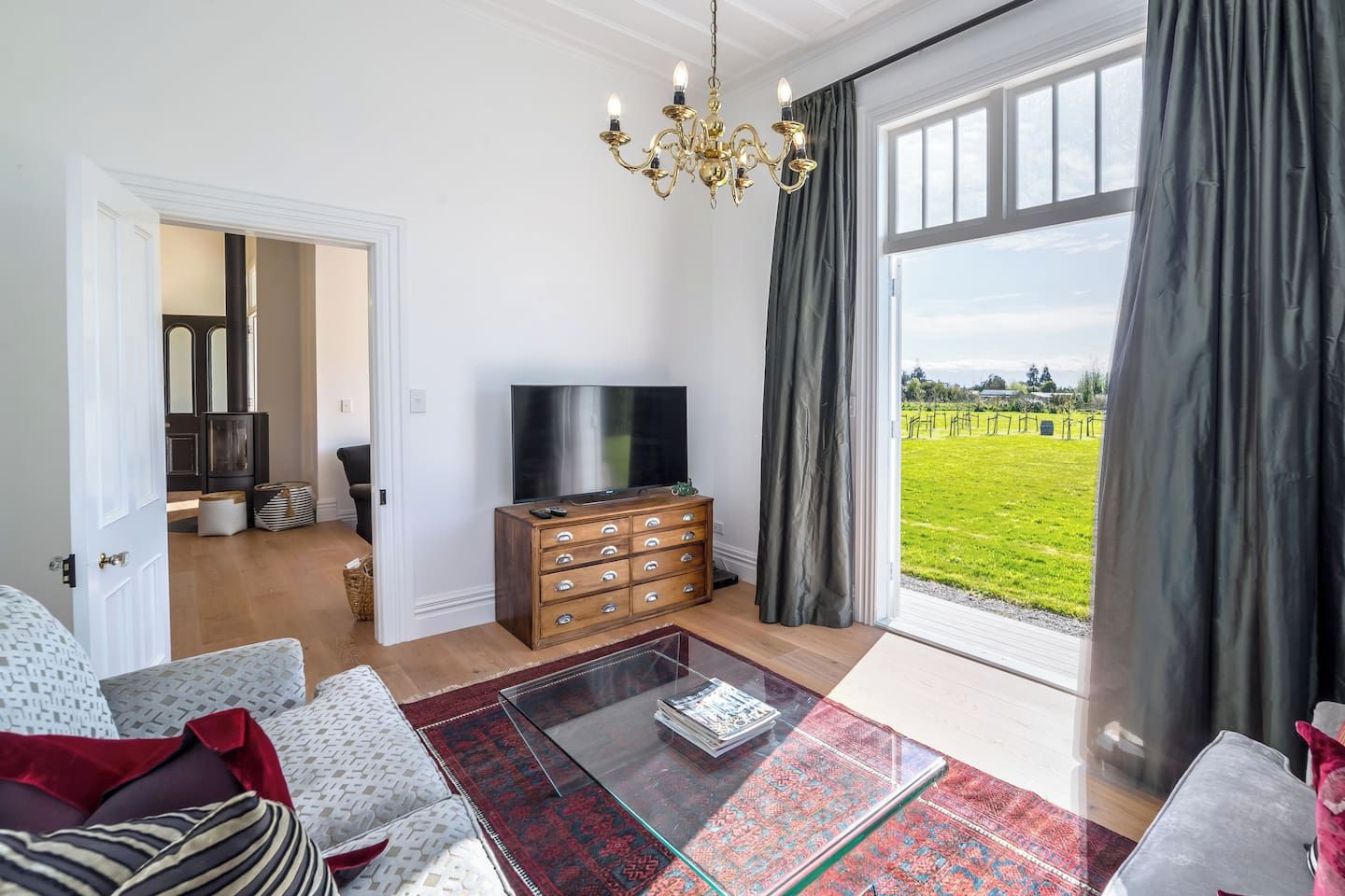 A living room with a television, a glass coffee table, and a view of a green field through large windows.