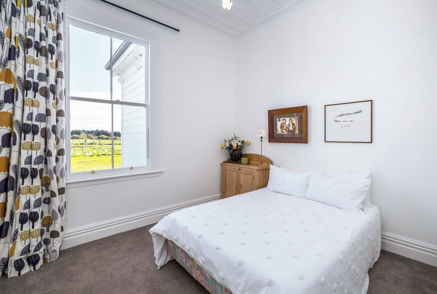 A bedroom with a large bed, a window with curtains, a wooden cabinet, and two framed pictures on the wall.