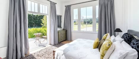 A bedroom with a large bed, grey curtains, a chandelier, and a view of greenery outside.