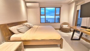 A modern bedroom with a wooden bed, a view of the sea through a large window, a flat-screen TV, and a ceiling with blue LED lighting.