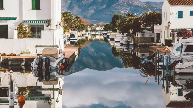 Un port de plaisance où des bateaux sont amarrés le long d’un canal paisible, entouré de bâtiments blancs aux volets verts, avec une chaîne de montagnes en arrière-plan.