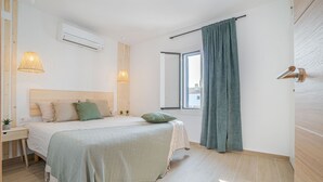 Une chambre à coucher comprenant un lit, des tables de chevet, un climatiseur, une fenêtre avec des rideaux et un plancher en bois.