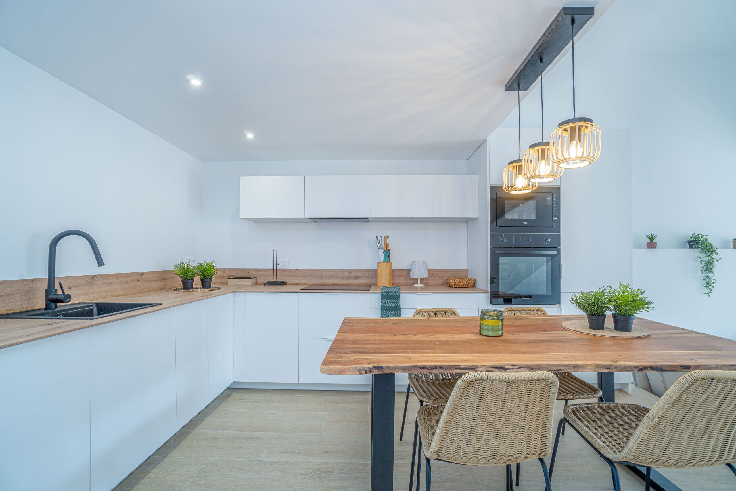 Een moderne keuken met witte kasten, een houten eettafel, rieten stoelen en hanglampen.