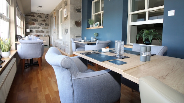 A modern dining area with a long table, grey upholstered chairs, and a stone wall. - Temple View Hotel (Isle of North Uist)