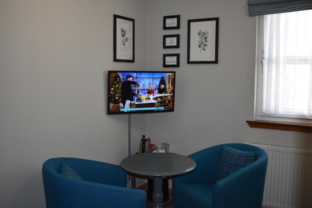 A room with a television mounted on the wall, two blue armchairs, a small round table, and framed botanical prints.