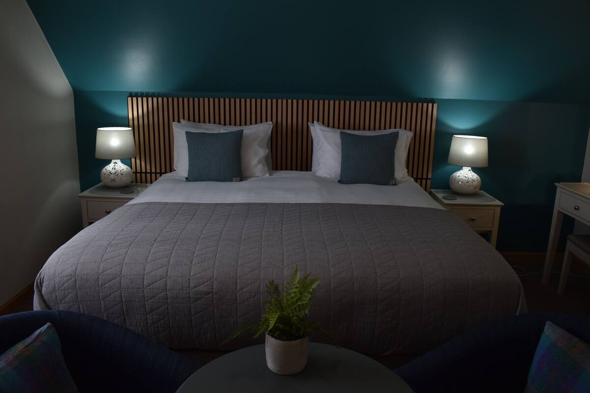 A neatly made bed with a grey quilt, two bedside lamps, and a potted plant on a round table.