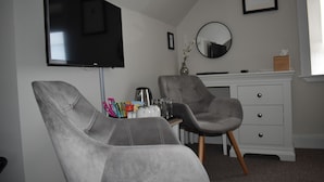 A modern living room with a flat-screen TV, two grey armchairs, a coffee table with various items, a white dresser with drawers, and a round mirror on the wall. - Temple View Hotel (Isle of North Uist)