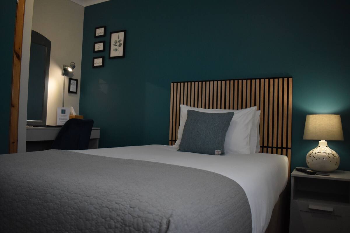A neatly made bed with a grey blanket, a bedside lamp, and a nightstand with a remote control.