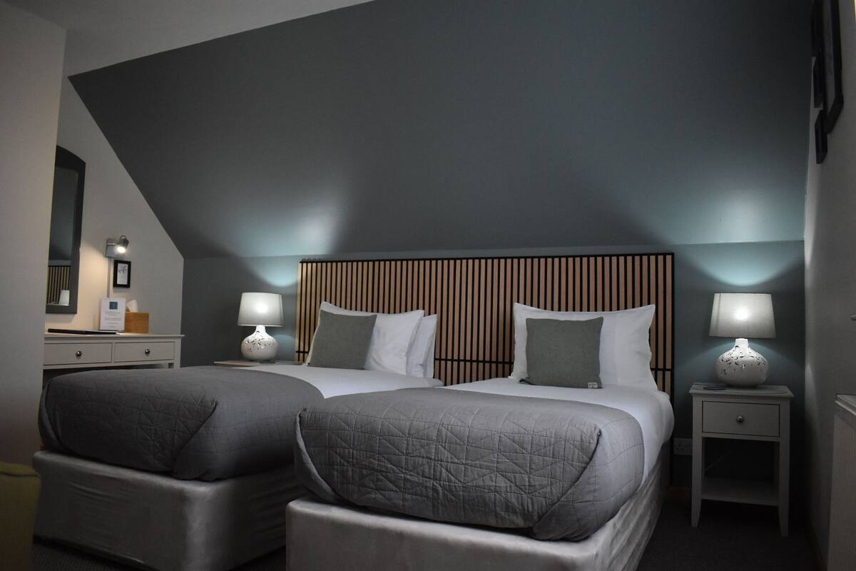A modern hotel room with two beds, a wooden headboard, bedside lamps, and a small nightstand.
