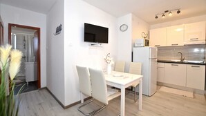 A modern kitchen with white cabinets, a built-in oven, and a refrigerator. There is a dining area with a white table and chairs, and a flat-screen TV mounted on the wall.