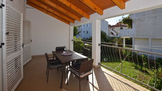 An outdoor patio with a wooden table and chairs, a white door, and a view of buildings and greenery.