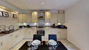 A modern kitchen with white cabinets, a dining table set with plates and glasses, and a coffee machine. - Orange Apartments - 150e Rusbislaw Mansions (Aberdeen)