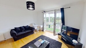 Una sala de estar con un sofá negro, un televisor de pantalla plana, una mesa de centro con un libro y un puerta corrediza de vidrio que da a un balcón.