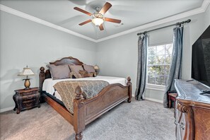 A bedroom with a wooden bed, a ceiling fan, a nightstand, a lamp, a window with curtains, and a dresser.