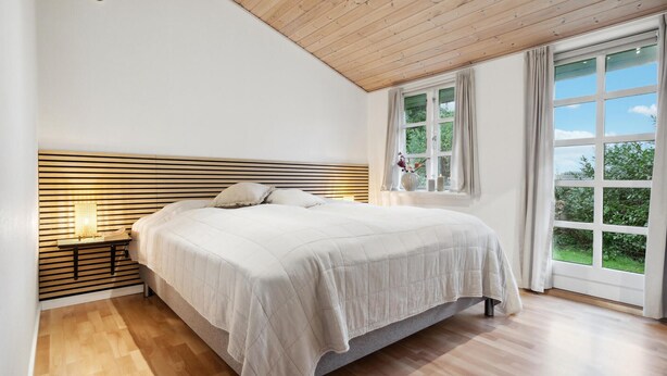 Una camera da letto con testiera in legno a listelli, un letto grande con copriletto bianco, un comodino con lampada e una finestra vista verde.