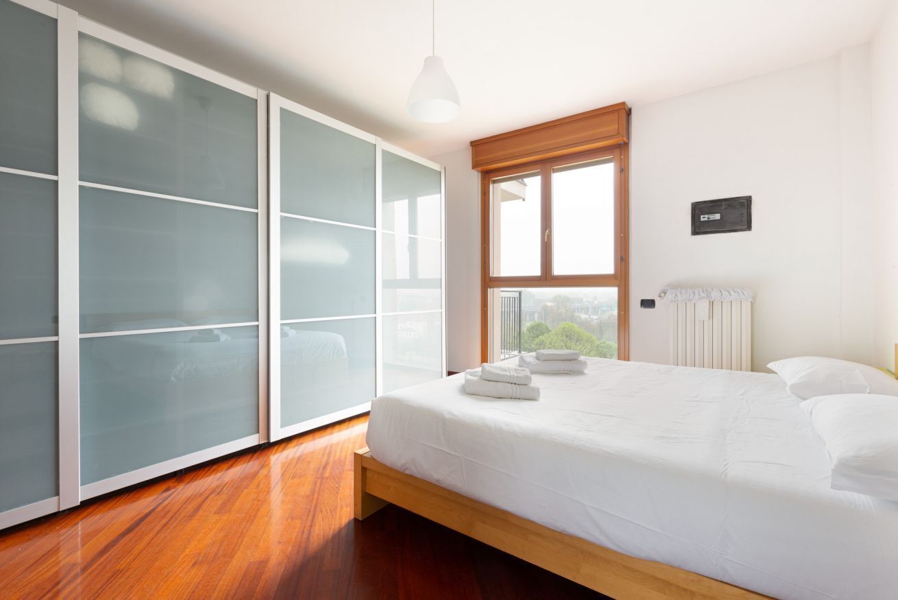Une chambre à coucher avec un lit, une grande fenêtre, un radiateur et une armoire aux portes en verre dépoli.