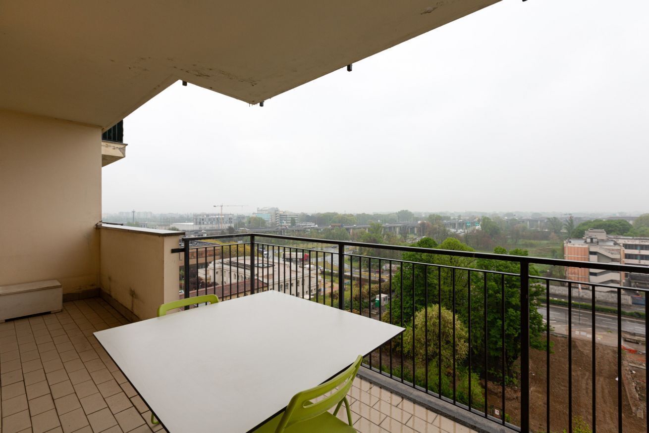 Ein Balkon mit einem weißen Tisch und grünen Stühlen, mit Blick auf eine Stadtlandschaft aus Gebäuden und Grünflächen.