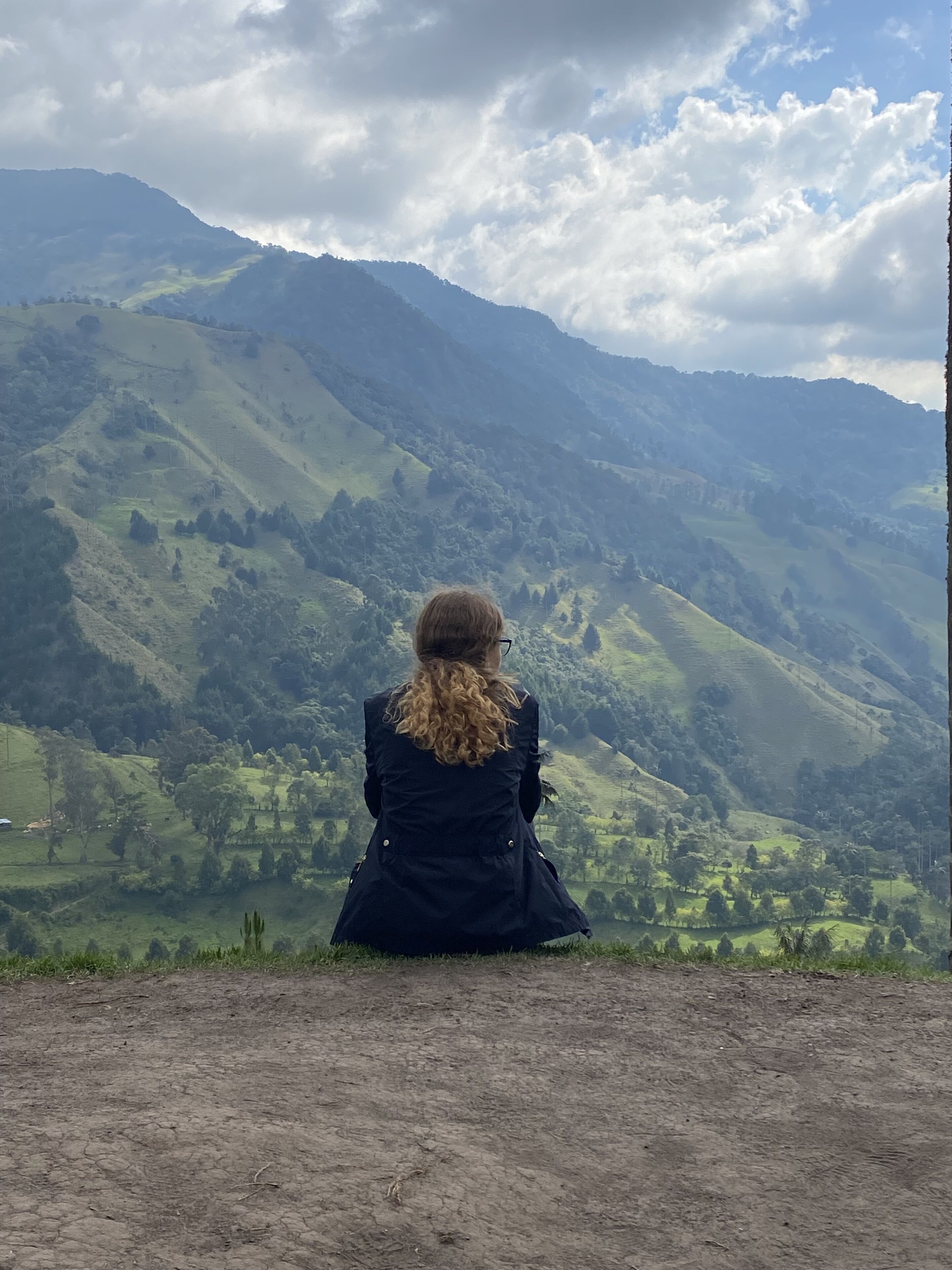 A person sitting on a dirt path overlooking a valley with hills and trees.