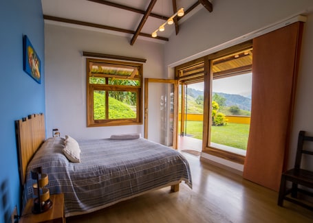 A bedroom with a large bed, wooden headboard, a bedside table, and a nightstand. There is a window with a view of greenery and mountains.